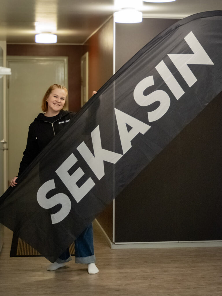 kuvassa punahiuksinen nainen ja musta lippu jossa lukee valkoisella sekasin