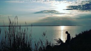 kuvassa merta, utuinen aurinko ja siluetti lapsesta lippis päässä. kuvassa etualla heinää