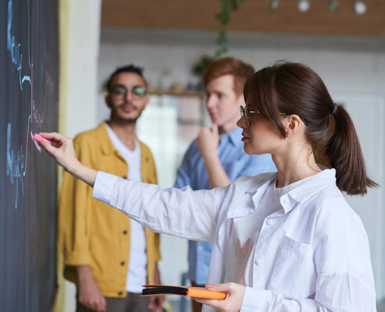 Naisoletettu piirtää taululle, taustalla kaksi ihmistä
